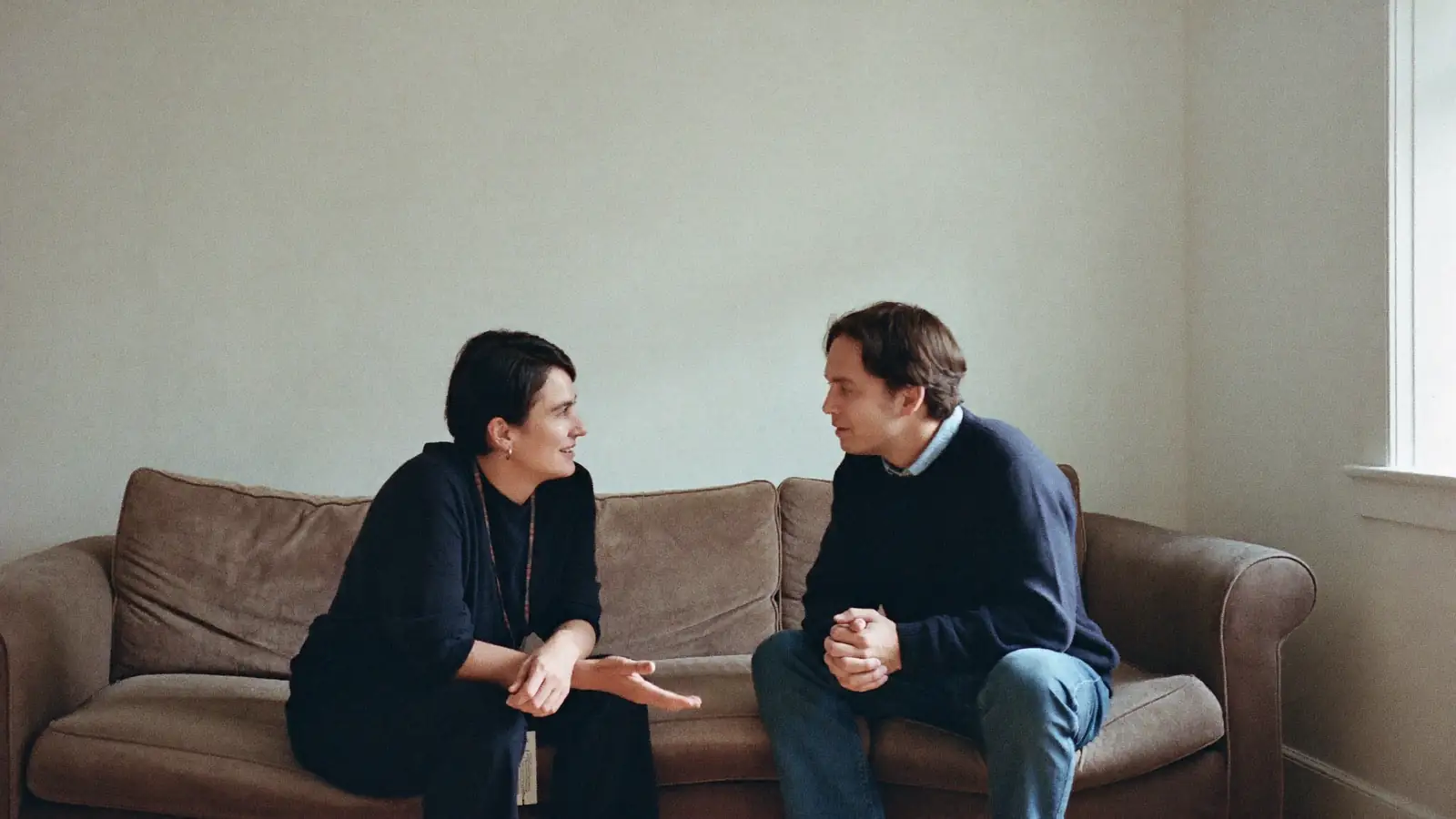 Two adults sitting together on a sofa in an ordinary living room, mid conversation.