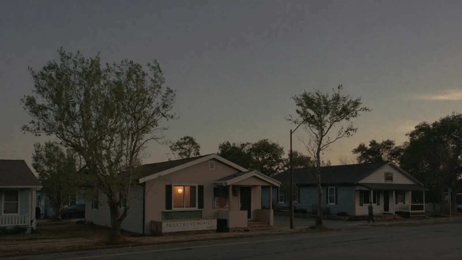 A quiet residential street at dusk with modest houses and warm light in one window.