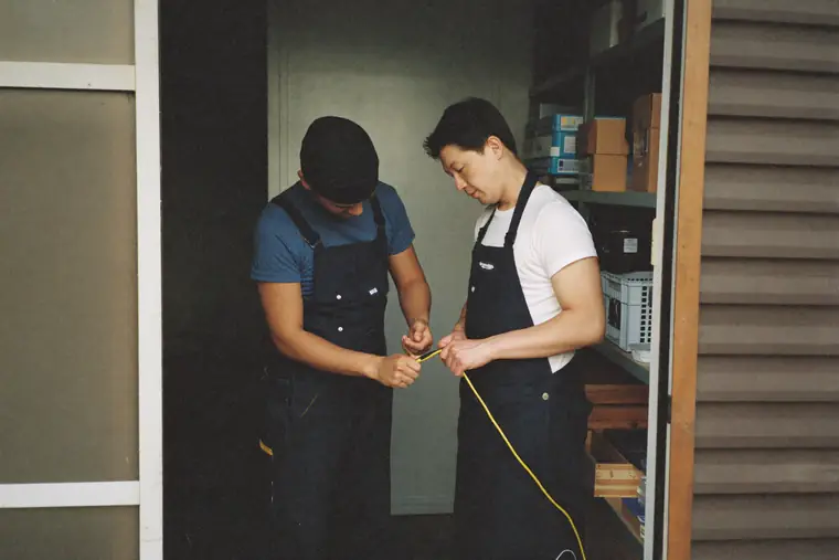Two adults in work clothes in a small workshop, one showing the other a task.