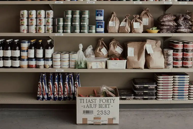 Shelves of tinned and dried food beside a crate of fresh vegetables at a community pantry.